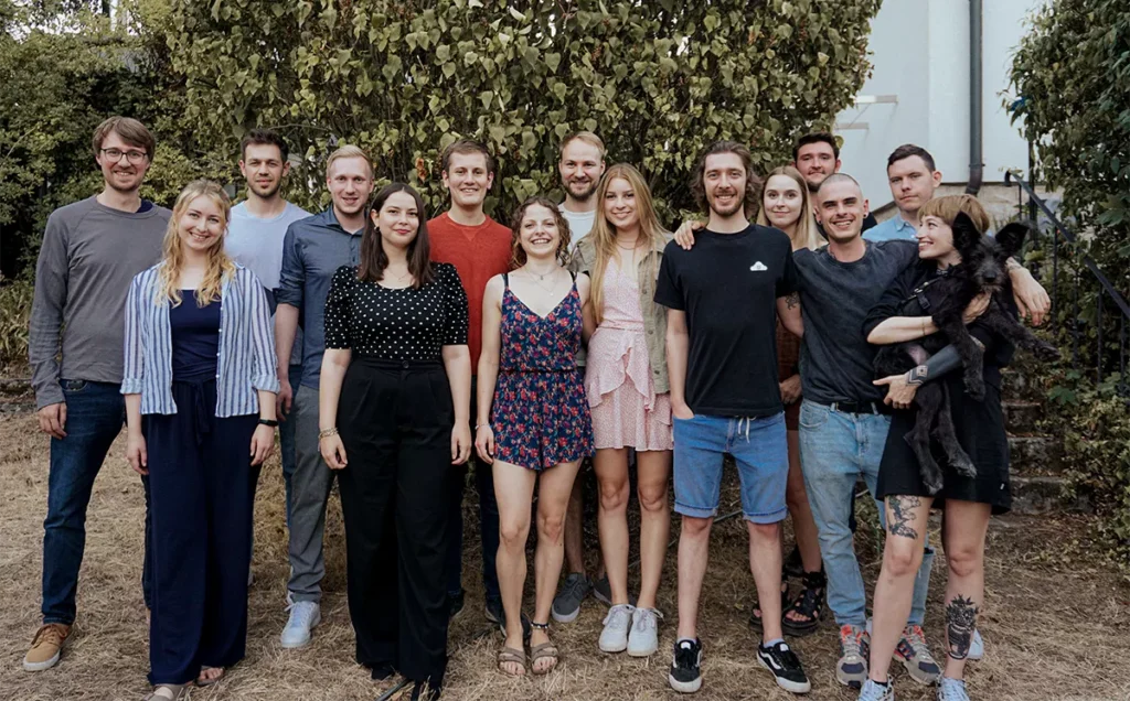 Il team di Eye-Able è in piedi davanti a uno sfondo verde. Si vedono 15 persone.